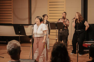 Caryl Loria Illana using crutches and singing into a mic on a stand, backed up by three singers