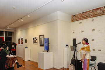 Caryl Loria Illana singing into a microphone on a stand, in the corner of a populated art gallery Caryl Loria Illana singing into a microphone on a stand, in the corner of a populated art gallery