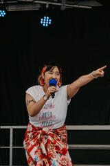 Caryl Loria Illana, in an &quot;Asian, queer, proud&quot; tee, singing into a microphone and pointing off camera