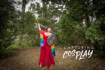 Caryl Loria Illana in the forest, wearing a rainbow cape, looking up with a sword and shield